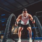 Focused man in white tank top performing battle rope exercise in a modern gym.