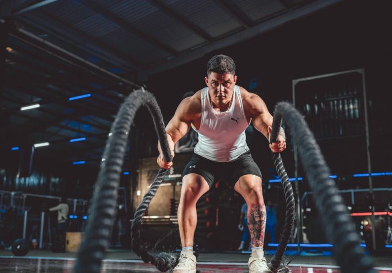 Focused man in white tank top performing battle rope exercise in a modern gym.